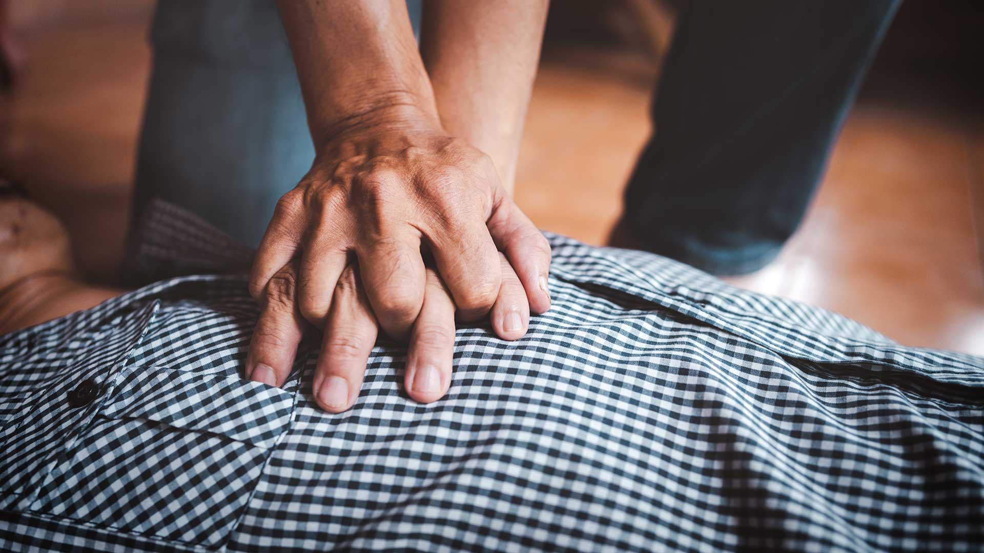Eine Hand führt eine Herzdruckmassage auf einem karierten Hemd aus, als Teil einer Ersten-Hilfe-Maßnahme.