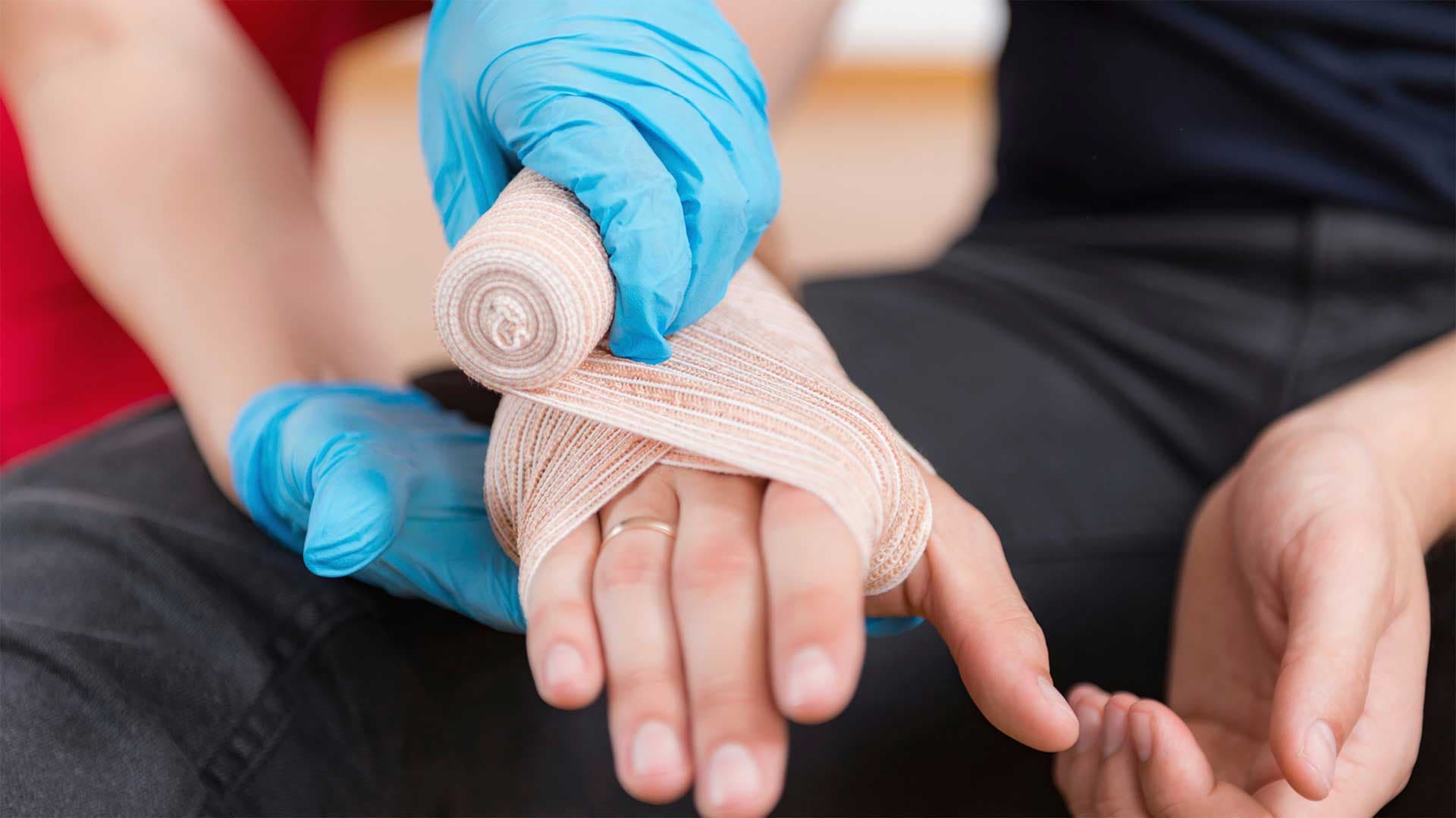 Ein Verband wird an der Hand einer Person angelegt, wobei der Helfer Handschuhe trägt, was auf die Einhaltung von Hygienehinweisen bei der Ersten Hilfe hinweist.