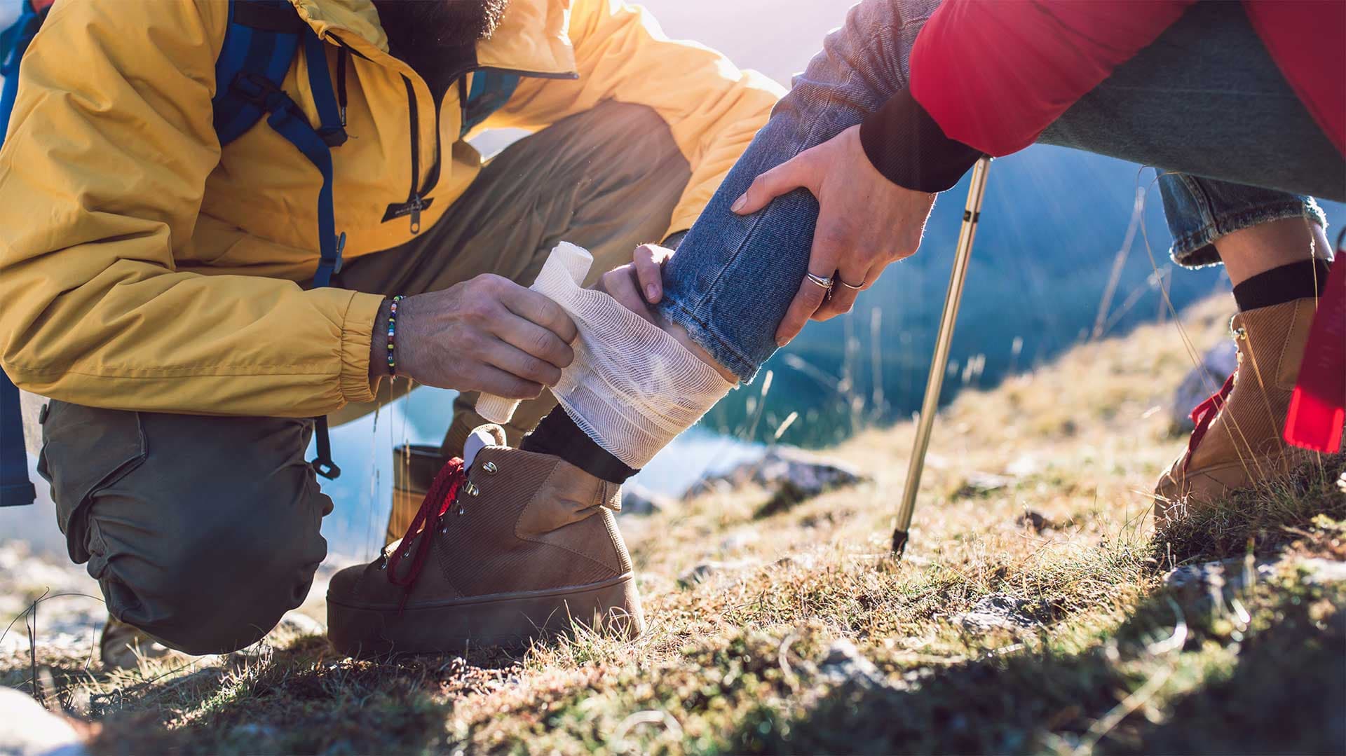 Ein Helfer wickelt einem verletzten Wanderer im Freien einen Verband um den Knöchel, beide tragen Wanderausrüstung.