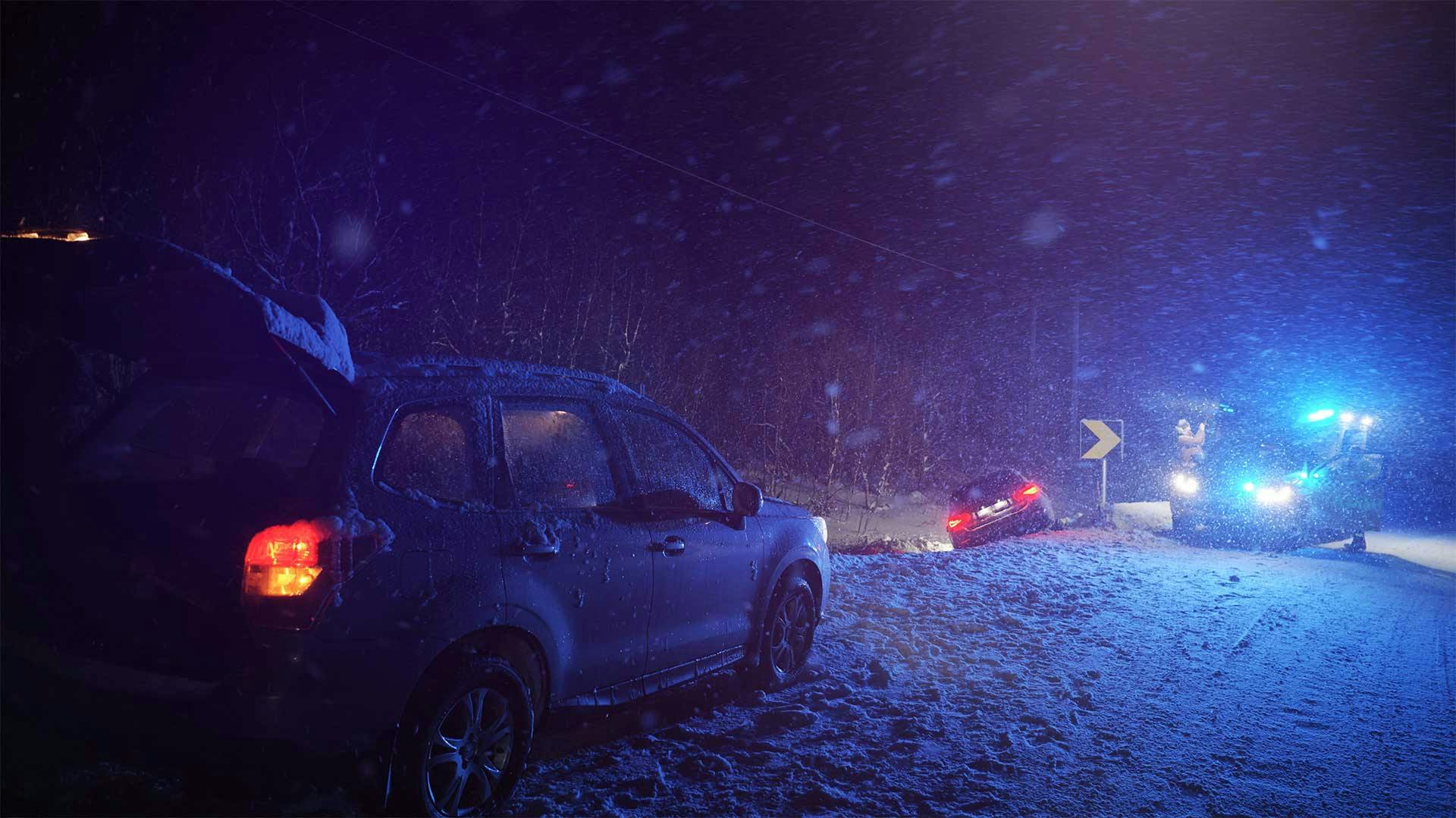 Zwei Autos im Schneesturm nachts an einem Unfallort, mit Blaulichtern der Rettungsfahrzeuge im Hintergrund.